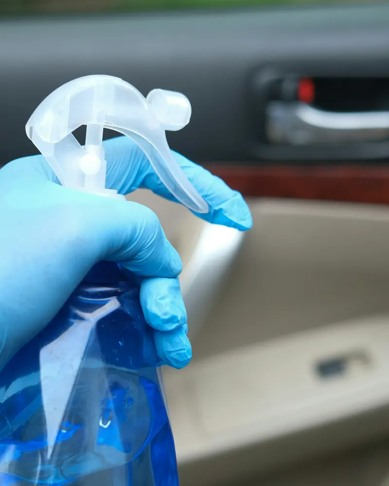 A gloved hand holding a spray bottle inside a car, by the door panel